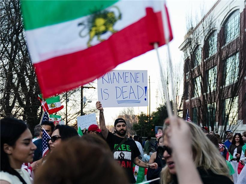 Mann mit Schild „Khamenei ist tot“ bei Kundgebung in Atlanta 2026, Menschen schwenken alte iranische Flaggen von 1979