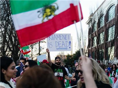 Mann mit Schild „Khamenei ist tot“ bei Kundgebung in Atlanta 2026, Menschen schwenken alte iranische Flaggen von 1979