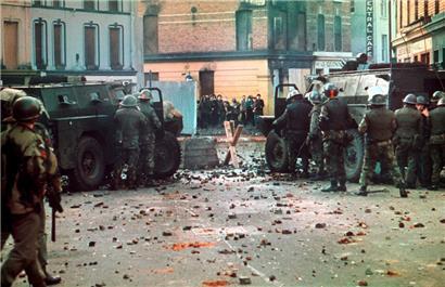 30.1.1972, Londonderry: Britische Soldaten mit Panzerwagen stehen auf einer mit Steinen übersäten Straße hinter einer Straßensperre
