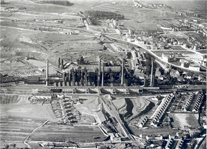 Blick auf das Hüttenwerk Esch-Terre Rouge und die vom Aachener Hütten-AV errichteten Arbeitersiedlungen: links die sieben Vierfachhäuser des Kazebierg, dann, neben dem Spielfeld der Jeunesse Esch, die 18 Doppelhäuser der rue des Mines, hinter dem Spielfeld die 17 Doppelhäuser der rue Renaudin. Im Hintergrund rechts eine Teilansicht der Stadtteile Brill, Uecht, Clair-Chêne und Auszéibréck, rue…
