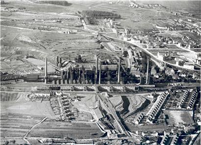 Blick auf das Hüttenwerk Esch-Terre Rouge und die vom Aachener Hütten-AV errichteten Arbeitersiedlungen: links die sieben Vierfachhäuser des Kazebierg, dann, neben dem Spielfeld der Jeunesse Esch, die 18 Doppelhäuser der rue des Mines, hinter dem Spielfeld die 17 Doppelhäuser der rue Renaudin. Im Hintergrund rechts eine Teilansicht der Stadtteile Brill, Uecht, Clair-Chêne und Auszéibréck, rue…
