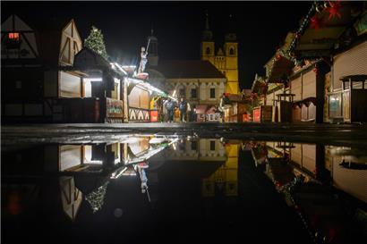 Buden des Weihnachtsmarktes, das Rathaus und die Johanniskirche spiegeln sich in einer Regenpfütze
