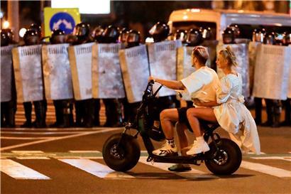 Ein Mann und eine Frau fahren auf einem Roller vor einer Reihe von Polizeibeamten, die mit Schutzschildern eine Straße blockieren. Nach der belarussischen Präsidentschaftswahl ist es zu blutigen Zusammenstößen von Polizei und Demonstranten gekommen. 

