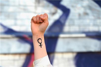Eine Frau hebt ihre Faust in die Höhe. Auf ihrem Arm ist das Venussymbol zu sehen, das für das weibliche Geschlecht und Frauenrechte steht.
