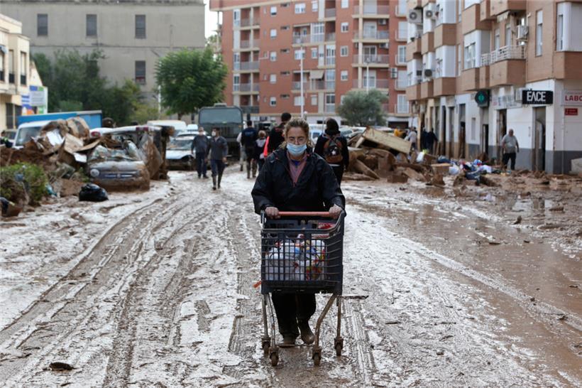 Eine Frau mit Mundschutz schiebt einen Supermarktwagen mit Lebensmitteln durch eine mit Schlamm bedeckte Straße in Paiporta bei Valencia in Spanien
