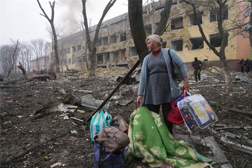 Eine Frau steht mit ihrem Gepäck vor einer durch einen Angriff beschädigten Geburtsklinik in Mariupol
