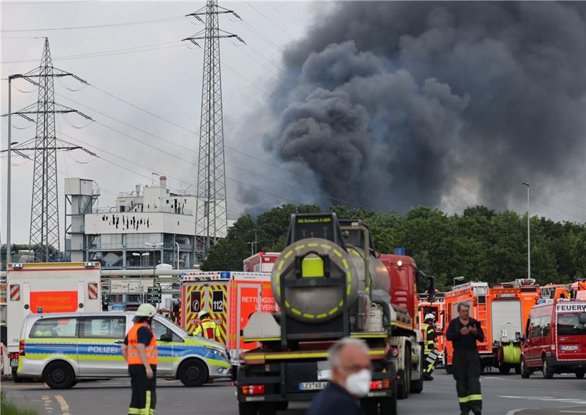 Einsatzfahrzeuge der Feuerwehr, Rettungsdienste und Polizei stehen unweit einer Zufahrt zum Chempark über dem eine dunkle Rauchwolke aufsteigt. 
