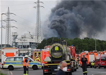 Einsatzfahrzeuge der Feuerwehr, Rettungsdienste und Polizei stehen unweit einer Zufahrt zum Chempark über dem eine dunkle Rauchwolke aufsteigt. 

