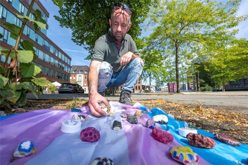 Felix Adrian Schäper vom Verein Trans*Inter*-Münster kniet vor einer Fahne mit Gedenksteinen, die Menschen auf dem Hafenplatz ausgelegt haben
