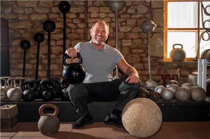 Georges Christen hockt in seiner Trainingsscheune vor historischen Hanteln und Gewichten
