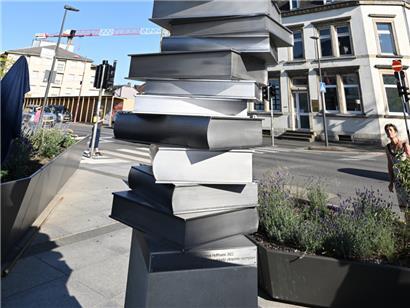 Im Auftrag der Gemeinde Petingen hat die Künstlerin Florence Hoffmann mit mehreren Partnern im Team eine monumentale Säulen-Skulptur in Form überdimensionaler, unregelmäßig aufgeschichteter Bücher aus rostfreiem Stahl geschaffen
