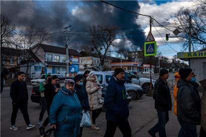In einem Ort knapp außerhalb von Kiew stehen die Menschen Schlange vor dem Supermarkt, im Hintergrund steigt der schwarze Rauch einer Explosion in den Himmel
