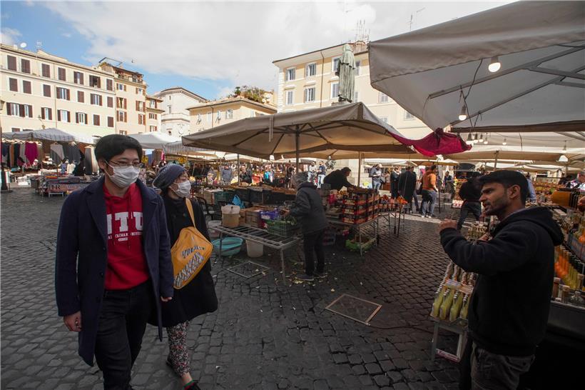 Italien, Rom: Zwei Passanten mit Mundschutz laufen während einem Marktes auf dem Platz «Campo de‘ Fiori» an einem Verkäufer vorbei. Die Zahl der Coronavirus-Infizierten und der Toten in Italien steigt trotz umfangreicher Gegenmaßnahmen stetig an. 
