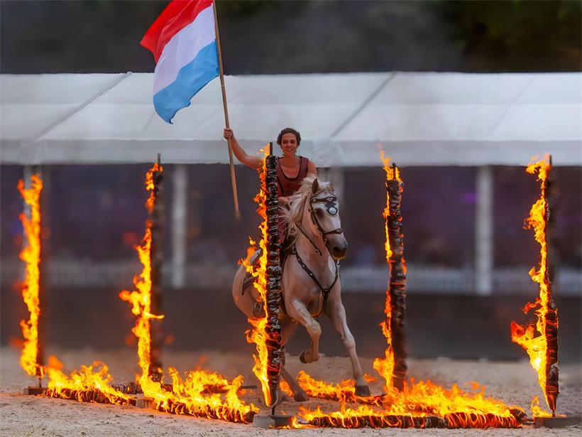 Jamie schwenkt stolz die luxemburgische Flagge, während Carbono mutig durch Flammen galoppiert, Action und Patriotismus