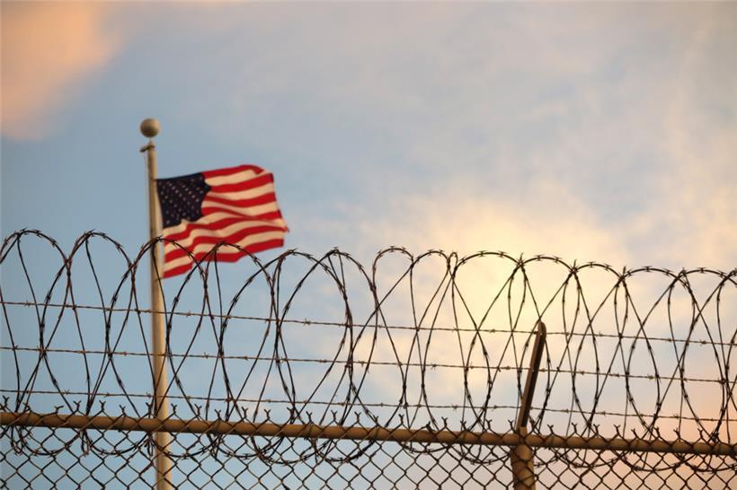 Kuba, Guantanamo Bay: Eine US-amerikanische Flagge weht hinter einem Stacheldrahtzaun im Wind.
