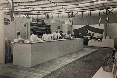 Luxemburgischer Stand auf der Messe in Léopoldville, dem heutigen Kinshasa, im Jahr 1951, im Hintergrund die Flagge Luxemburgs neben jener des Belgisch‑Kongo sowie ein Porträt der Großherzogin Charlotte in der Mitte