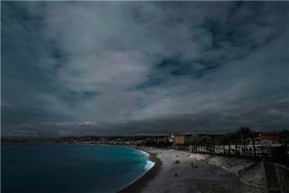 Menschen sitzen am Strand oder spazieren entlang der berühmten „Promenade des Anglais“ unter einem bewölkten Himmel in Nizza
