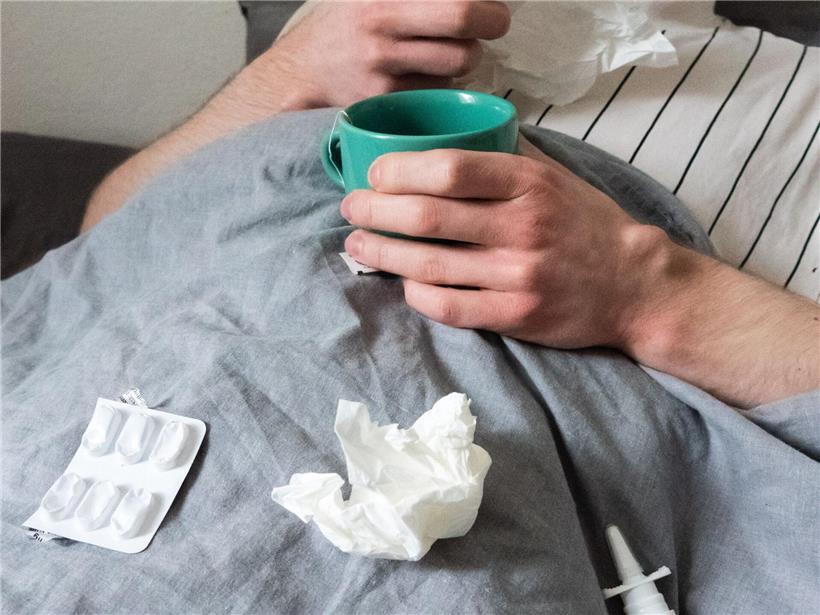 Mann im Bett mit Erkältung, umgeben von Taschentüchern, Teetasse, Nasenspray und Tabletten zur Genesung