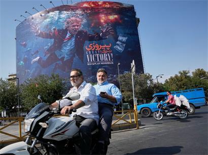 Motorradfahrer fahren an einer Plakatwand vorbei, auf der US-Präsident Trump unter Wasser inmitten von Explosionen zu sehen ist, begleitet von der Aufschrift „Großer Sieg! Straße von Hormus“