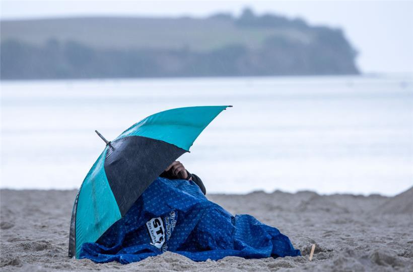 Urlauber sitzen Anfang August bei kräftigem Regen unter einem Schirm am Strand in Norddeutschland
