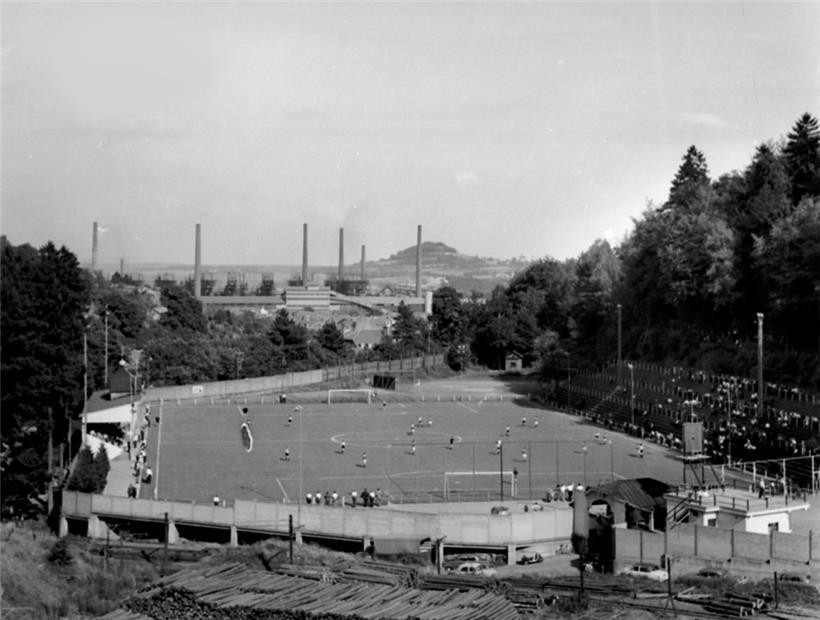 Vue sur le Stade du Thillenberg en 1958, avec la tribune et les gradins et le cadre boisé. A l’avant-plan, le bois stocké pour la mine du Thillenberg. En arrière-plan la ligne des dix hauts-fourneaux de l’usine de Differdange et la Zolverknapp.
