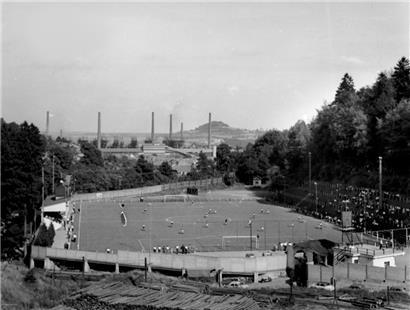 Vue sur le Stade du Thillenberg en 1958, avec la tribune et les gradins et le cadre boisé. A l’avant-plan, le bois stocké pour la mine du Thillenberg. En arrière-plan la ligne des dix hauts-fourneaux de l’usine de Differdange et la Zolverknapp.
