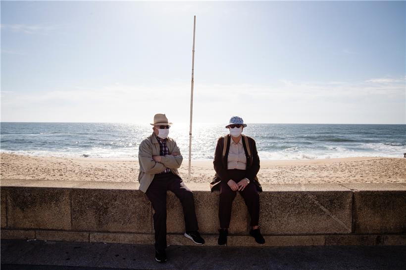 Zwei ältere Männer sitzen Anfang Mai mit Mundschutz am Strand von Espinho – bald dürften dahinter wieder Liegen und Schirme stehen
