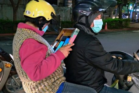 Eine Frau mit Mundschutz sitzt in Taiwan als Beifahrerin eines Mannes, der ebenfalls einen Mundschutz trägt, auf einem Motorrad und liest auf einem Smartphone und einem Tablett.