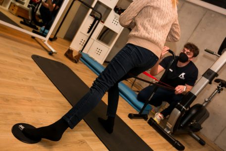 Diese Übung nutzt Bewegungsabläufe aus dem Langlauf, eine Technik, wie sie beispielsweise beim Skating im Langlauf eingesetzt wird. Physiotherapeut Yannick Zenner hilft mit einem Band – zu Hause klappt es auch mit einem Schal, damit das Knie beim schwungvollen Schritt nach vorn, nicht seitlich abknickt, sondern nach vorne gerichtet bleibt