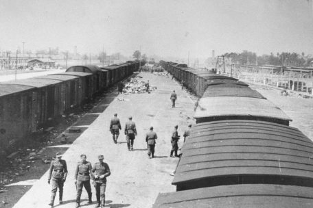 SS-Männer gehen entlang der neuen Ankunftsrampe in Auschwitz-Birkenau. Die Rampe wurde verlängert, damit die Todeszüge ab Frühjahr 1944 direkt bis zu den Vernichtungsanlagen heranfahren konnten. Die Krematorien II und III (erkennbar an den Schornsteinen) sind im Hintergrund rechts und links von den Güterwaggons zu sehen. Foto von Bernhard Walter/Ernst Hofmann, Mai 1944.