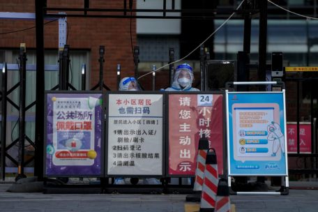 27.11.2022, China, Peking: Sicherheitskräfte in Schutzanzügen halten Wache in der Nähe von Hinweisschildern zur Corona-Prävention, die vor einem Wohnviertel in Peking ausgestellt sind